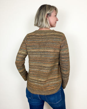 A woman with short blonde hair is shown from the back wearing a textured, striped knit sweater in earthy brown, beige, and muted green tones, paired with blue jeans, against a plain white wall.