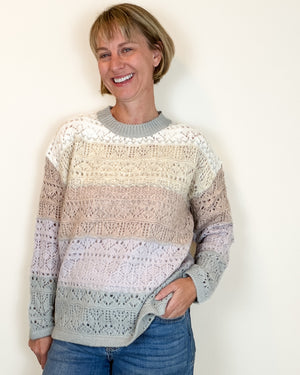 A smiling woman with short blonde hair wearing a striped, open-knit crewneck sweater in shades of cream, beige, pink, and light gray, paired with blue jeans, standing against a plain off-white wall.
