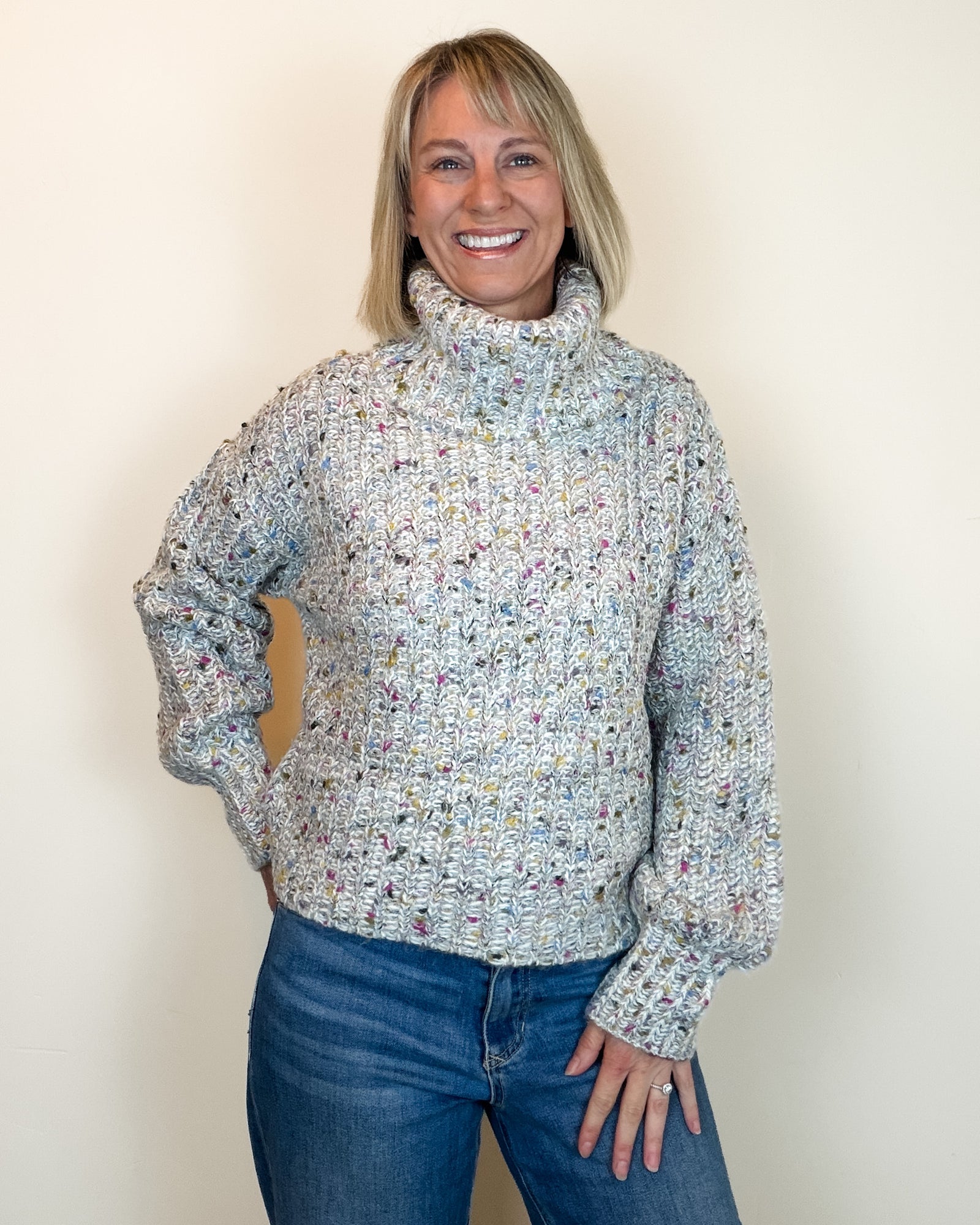A smiling blonde woman wearing a chunky knit turtleneck sweater with a multicolor variegated pattern, paired with blue jeans, standing against a plain off-white wall.