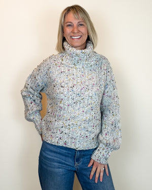 A smiling blonde woman wearing a chunky knit turtleneck sweater with a multicolor variegated pattern, paired with blue jeans, standing against a plain off-white wall.