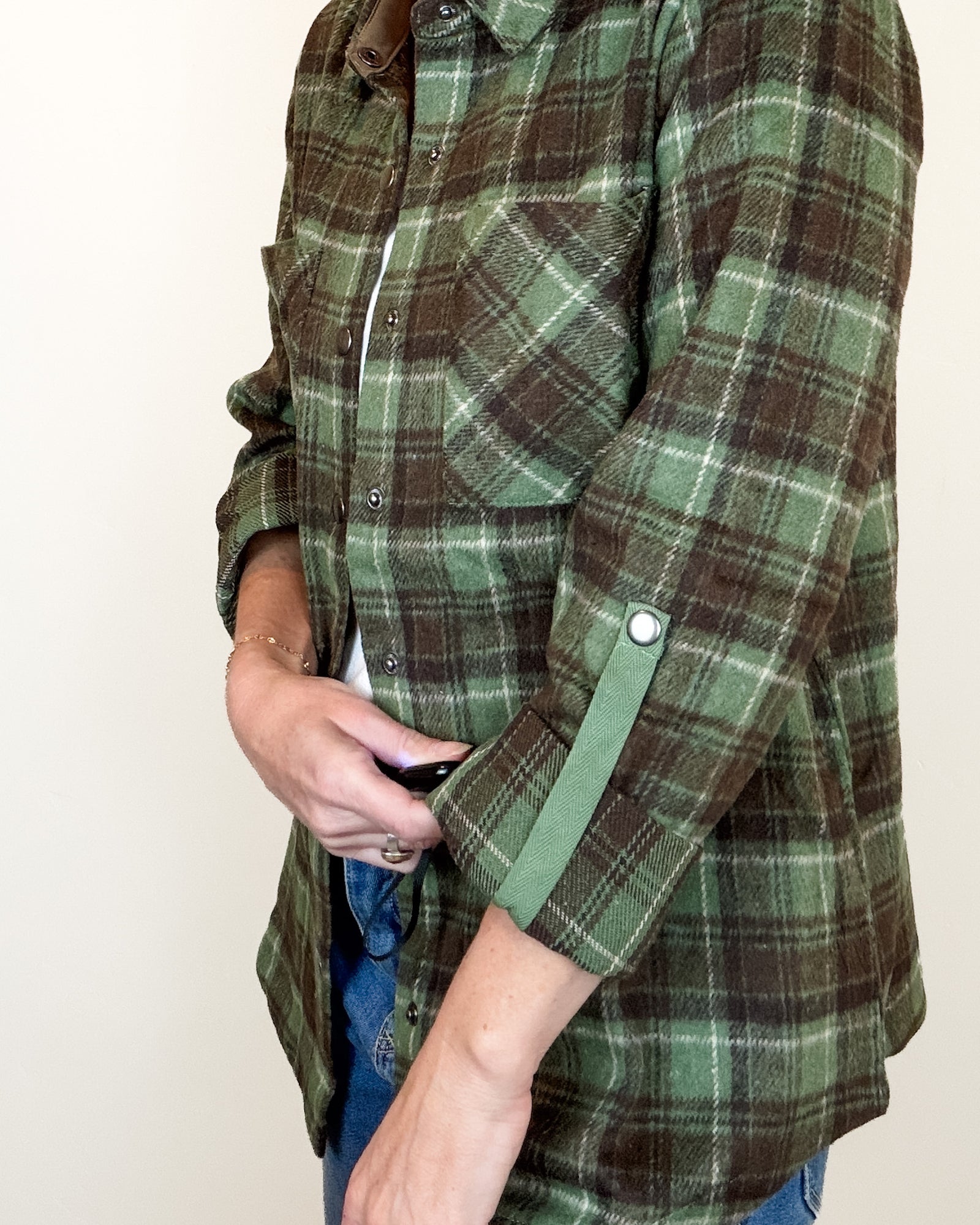 A woman modeling a green and brown plaid button-up flannel shirt over a white undershirt and blue jeans, standing against a plain off-white wall.