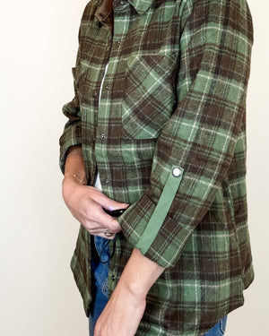 A person wearing a green and brown plaid button-up flannel shirt with blue jeans, shown from the chest down against a plain off-white background.