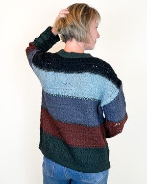 A woman shown from the back wearing a chunky, multi-colored striped knit sweater with blue, burgundy, and dark green horizontal bands, paired with blue jeans against a plain white background.