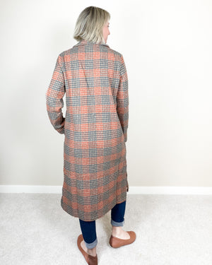 A woman viewed from behind wearing a long-sleeved, knee-length plaid coat in orange and gray over a dark blue dress, standing on a light gray carpet against a white wall.