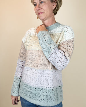 A woman wearing a long-sleeve knit sweater with horizontal color-blocked panels in cream, beige, light blue, and white, paired with blue jeans against a solid beige background.
