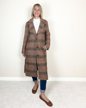 A woman modeling a long brown and beige plaid coat over a white turtleneck, blue jeans, and brown flats, standing against a plain off-white wall and light carpet.