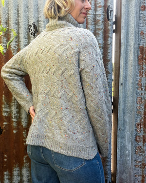 A woman wearing a light gray cable-knit turtleneck sweater with multicolored flecks, paired with blue jeans, standing in front of a rustic corrugated metal door.