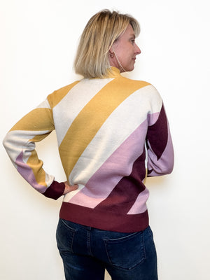 A woman with blonde hair is shown from the back wearing a long-sleeve mock-neck sweater with bold diagonal color-blocked stripes in mustard yellow, cream, light pink, and deep burgundy, paired with dark blue jeans against a plain white background.