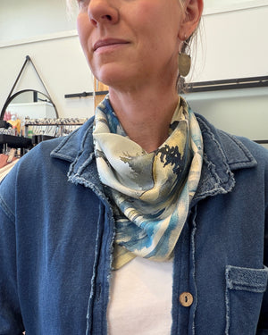 A close-up lifestyle shot of a woman wearing a blue denim jacket over a white t-shirt, with a patterned silk scarf in blue, beige, and white draped around her neck. She is wearing large gold-toned earrings in a retail store setting.