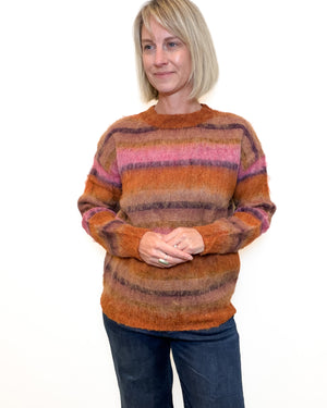A woman with blonde hair wearing a fuzzy, striped crewneck sweater in shades of orange, brown, and pink, paired with dark blue jeans, standing against a plain white background.