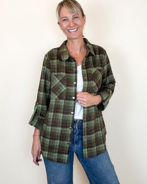 A woman modeling a green and brown plaid button-up flannel shirt over a white undershirt and blue jeans, standing against a plain off-white wall.
