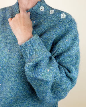 A close-up of a person wearing a textured blue knit sweater with three decorative silver buttons at the collar and small scattered sequins.