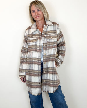 A woman modeling a long-sleeve, fringed plaid button-up shirt jacket in beige, brown, and white, paired with blue jeans against a plain white wall.