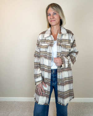 A woman modeling a long-sleeve, fringed plaid coat over a white top and blue jeans, standing against a plain beige wall.
