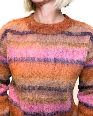 A close-up of a person wearing a fuzzy striped sweater with horizontal bands of orange, pink, purple, and brown on a plain light background.