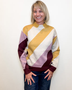 A smiling blonde woman wearing a color-block knit sweater with diagonal stripes in mustard yellow, cream, dusty lavender, and deep burgundy, paired with dark blue jeans, standing against a plain off-white wall.