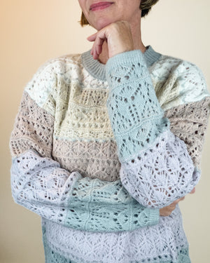 A person wearing a long-sleeve open-knit sweater with horizontal color-blocked panels in pale blue, cream, and taupe, posed against a plain beige background.