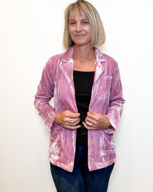 A woman with blonde hair wearing a pink and white marbled textured jacket over a black top and blue jeans, standing against a plain white wall.