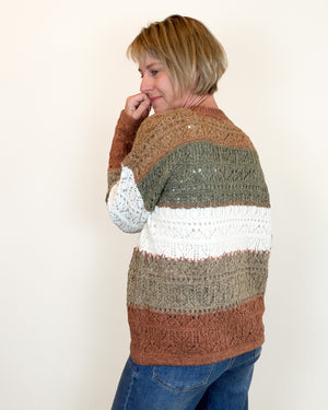 A woman wearing a chunky knit striped sweater with horizontal bands of rust brown, olive green, and white, paired with blue jeans, photographed against a plain off-white background.
