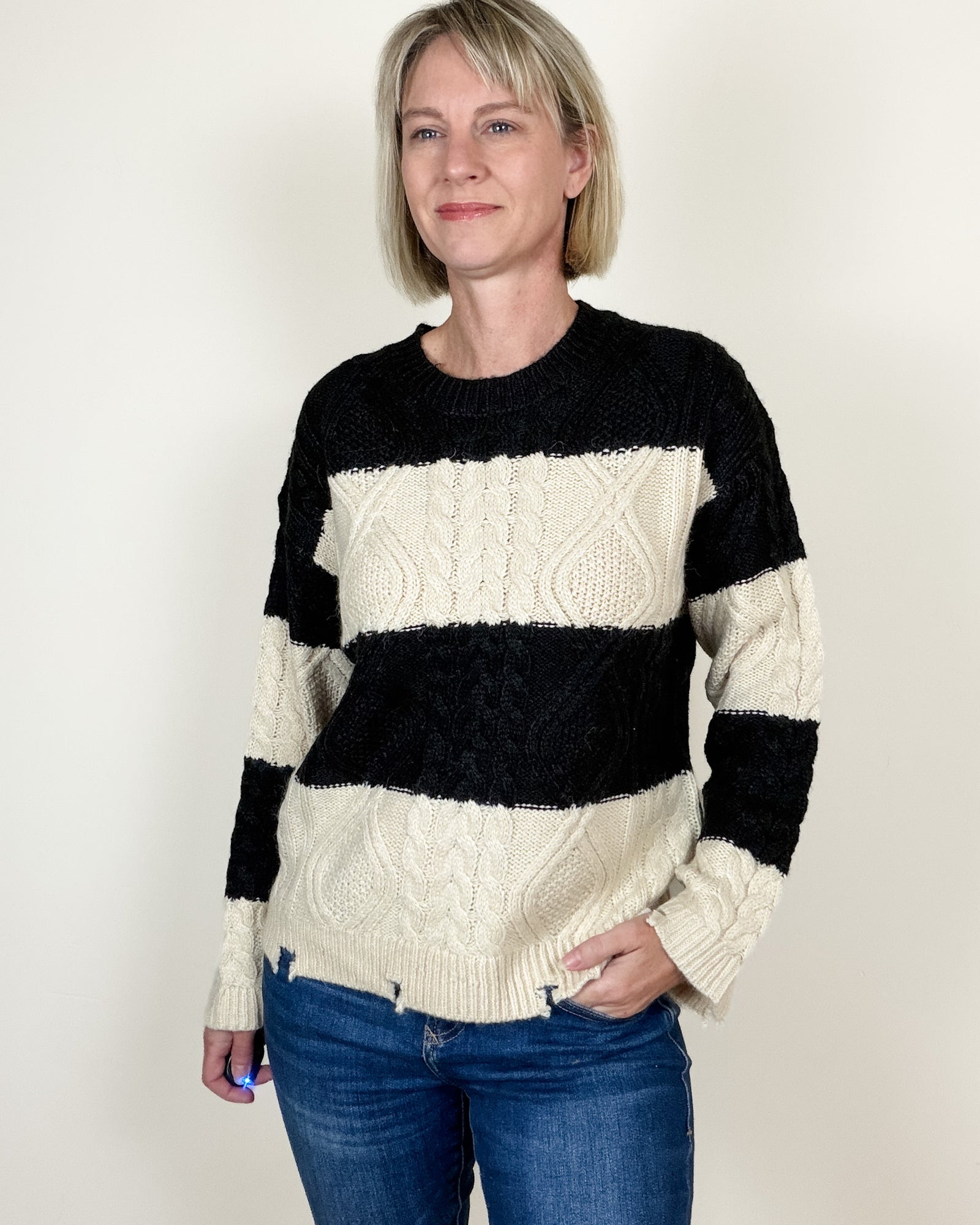 A smiling woman with short blonde hair wearing a black and cream cable-knit sweater and blue jeans, standing against a plain off-white background.