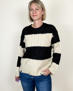 A woman wearing a black and cream striped cable-knit sweater with blue jeans, standing against a plain off-white background.