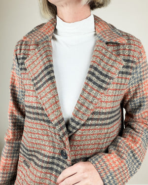 A close-up of a person wearing a textured plaid blazer in orange, gray, and beige tones over a white turtleneck.