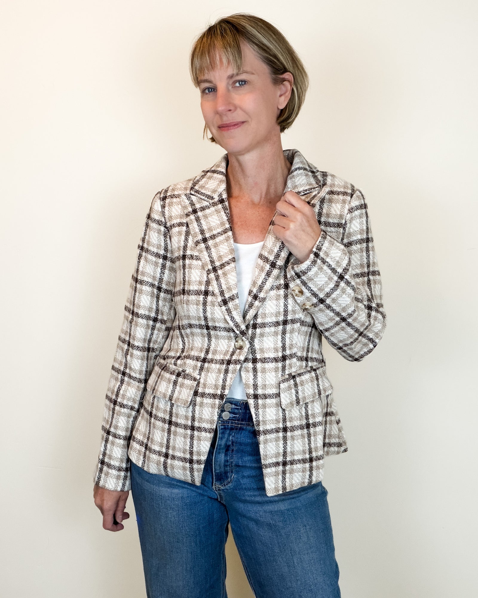 A woman modeling a beige and brown plaid blazer over a white top and blue jeans, standing against a plain off-white background.