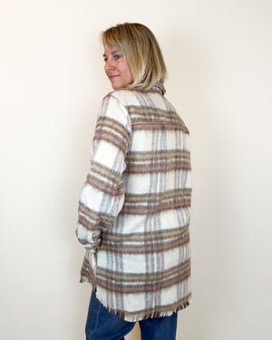 A woman with blonde hair is shown from the back wearing a loose, fringed plaid cardigan in off-white, brown, and gray, paired with blue jeans, against a plain off-white wall.