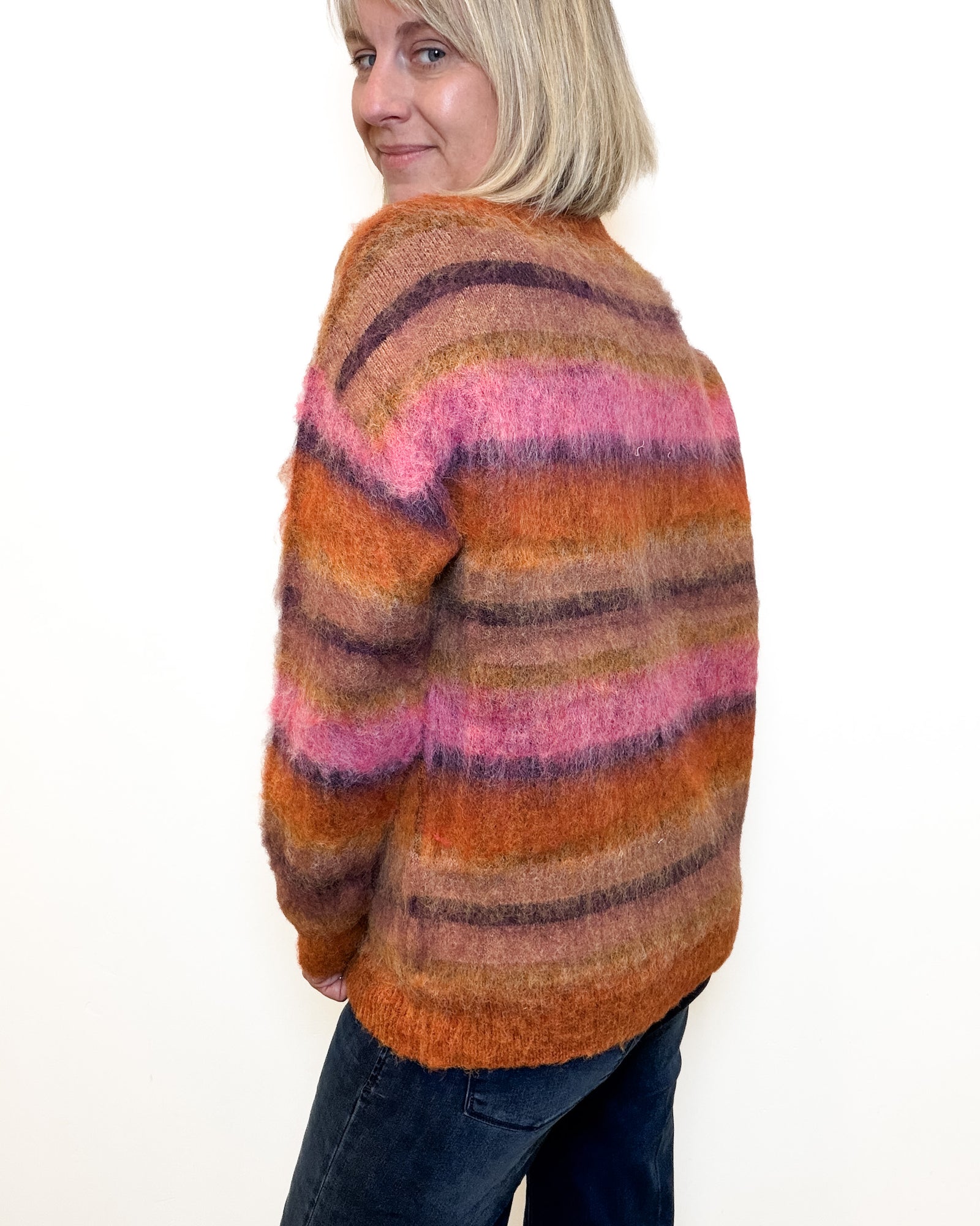 A woman with blonde hair wearing a fuzzy, striped crewneck sweater in shades of orange, brown, and pink, paired with dark blue jeans, standing against a plain white background.