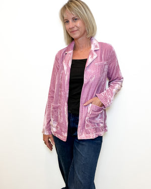 A woman modeling a pink textured velvet jacket with a black top and blue jeans, standing against a plain white background.