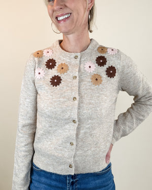 A woman wearing a light heather gray knit cardigan with crocheted flower appliqués in brown, tan, and pale pink across the chest, paired with blue jeans against a plain beige background.