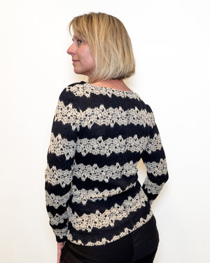 A woman with blonde hair is shown from the back wearing a black and cream knit sweater with horizontal lace-like patterns, set against a plain white background.