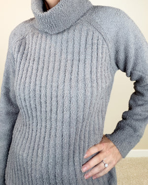 A close-up of a person wearing a light gray ribbed turtleneck sweater, showing the textured knit fabric and a hand resting on the hip.