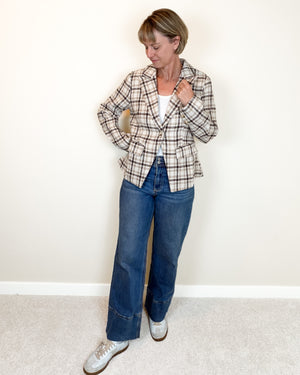 A woman modeling a beige and brown plaid blazer over a white top, paired with blue jeans and white sneakers, standing against a plain off-white wall.