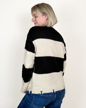 A woman with short blonde hair is shown from the back wearing a black and cream striped knit sweater and blue jeans against a plain white background.