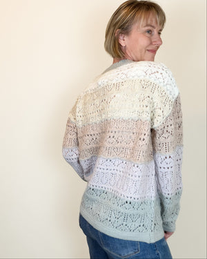 A woman wearing a long-sleeve, open-knit sweater with horizontal stripes in cream, beige, light brown, and pale blue, paired with blue jeans, shown from a side-back angle against a plain beige wall.