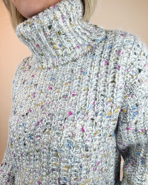 Close-up of a person wearing a chunky knit turtleneck sweater in a light gray and white variegated yarn with small multicolored flecks, against a plain beige background.