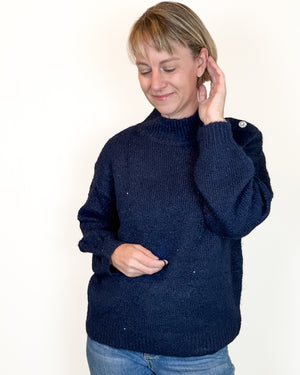 A woman wearing a dark navy blue ribbed knit sweater with a mock neck and buttoned shoulder detail, paired with blue jeans, standing against a plain off-white background.