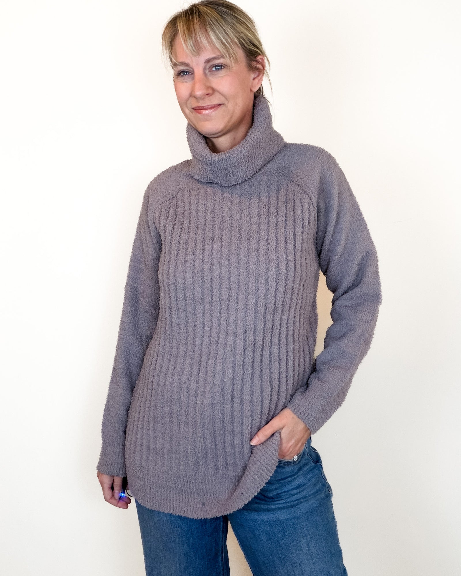 A woman wearing a chunky ribbed turtleneck sweater in a dusty mauve-gray color, paired with blue jeans, standing against a plain off-white background.
