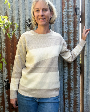 A woman with short blonde hair stands in front of a rustic corrugated metal gate, wearing a cream and beige striped knit sweater and blue jeans.
