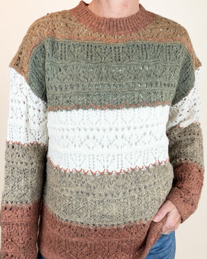 A person wearing a chunky knit crewneck sweater with horizontal color-blocked stripes in olive green, white, taupe, and rust brown, paired with blue jeans against a plain beige background.