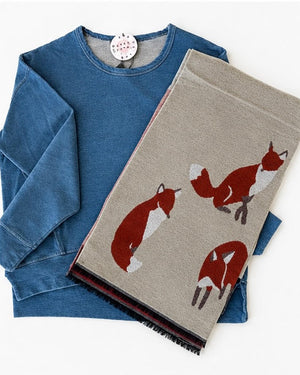 A blue long-sleeve top laid flat with a beige scarf draped over it. The scarf features three red and white fox illustrations and has black fringe along one edge.