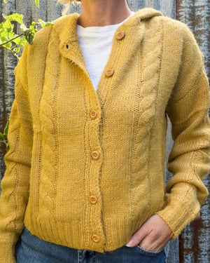 A person wearing a mustard yellow cable-knit cardigan with wooden buttons over a white t-shirt and blue jeans, standing against a rustic corrugated metal wall.