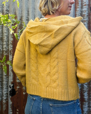 A person shown from the back wearing a mustard yellow cable-knit cropped hoodie with a hood, paired with blue jeans, standing against a rustic corrugated metal wall.