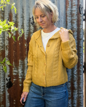 A woman wearing a mustard yellow cable-knit cardigan with a hood, styled over a white t-shirt and blue jeans, standing in front of a corrugated metal wall.