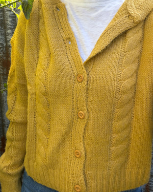 A close-up of a person wearing a mustard yellow cable-knit cardigan over a white top and blue jeans, with outdoor foliage and a fence visible in the background.