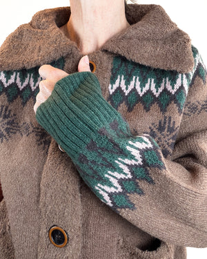 Close-up of a person wearing a brown knit coat with a green and white patterned knit collar and sleeve, holding the coat closed with one hand.
