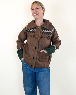 A woman wearing a brown fuzzy knit cardigan with a dark green and white geometric pattern across the chest and sleeves, paired with blue jeans, standing against a plain off-white background.