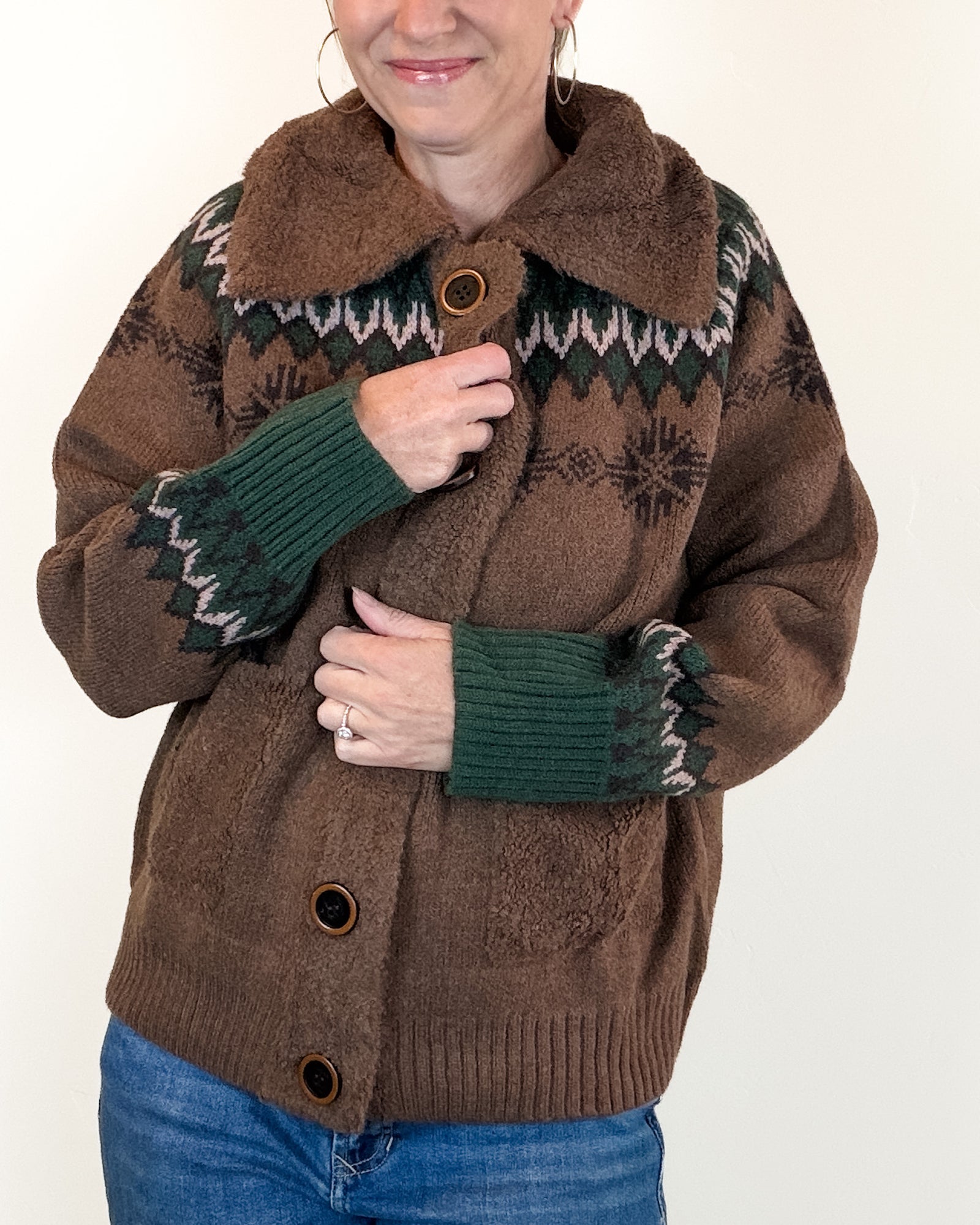 A woman wearing a brown fuzzy knit cardigan with a dark green and white geometric pattern, ribbed green cuffs, and brown buttons, paired with blue jeans against a plain white background.