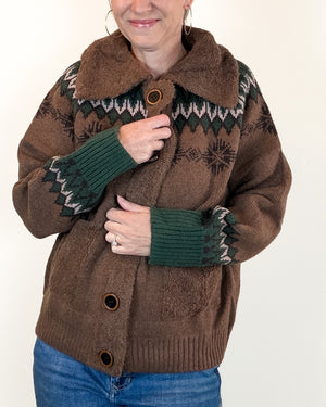 A woman wearing a brown fuzzy knit cardigan with a dark green and white geometric pattern, ribbed green cuffs, and brown buttons, paired with blue jeans against a plain white background.